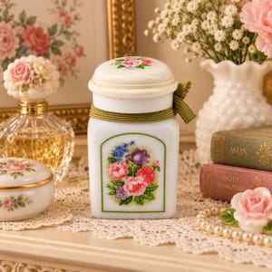 May include: A white ceramic jar with a lid, featuring a floral bouquet decal on the front, a green ribbon tied around the neck, and a matching floral design on the lid. The jar has a square base with rounded corners and a smooth, glossy finish.