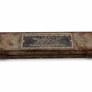 May include: A rectangular, weathered metal storage case with a dark, rusted finish. A black metal identification plate is riveted to the top, featuring white text: HANDLES; SOCKET WRENCH, TORQUE-INDICATING, SQUARE DRIVE, TYPE I, SIZE 1, RANGE 0-100 IN-LBS.