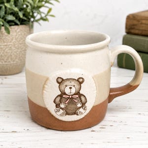 Puede incluir: Una taza de cerámica con cuerpo de color crema y beige moteado, con base y asa de color marrón. Un parche circular en relieve en la parte frontal muestra una ilustración de un oso de peluche marrón oscuro con una pajarita de color rosa claro.