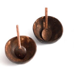 May include: Two handcrafted coconut bowls with matching spoons. The bowls are dark brown with a natural wood grain, and each spoon rests inside. The spoons have a long handle and a rounded scoop. The bowls and spoons are on a white background.
