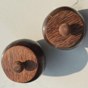 以下が含まれることがあります： 木製の蓋が付いた、丸い濃い茶色のココナッツ殻容器が2つ。各蓋には、小さな丸い木製のつまみが付いています。容器は明るい色の表面にあり、影ができています。