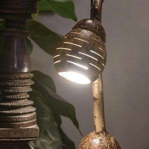 May include: A handcrafted table lamp with a unique design. The lamp features a coconut shell shade with horizontal cutouts, mounted on a wooden stem and base. The warm light shines through the openings, creating a cozy ambiance. The lamp is approximately 30 cm tall.