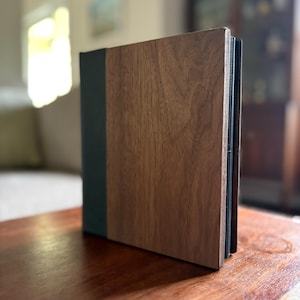 May include: A rectangular photo album featuring a front cover with a prominent wood grain texture in a medium brown tone, contrasted by a solid dark gray spine. The album is standing upright, showcasing its layered construction and clean edges.