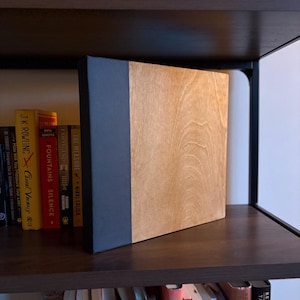 May include: A rectangular photo album or book with a natural light brown wood grain front cover and a solid matte black spine. The object is positioned upright, showcasing a clean, two-tone design with a smooth finish on both the wood and black sections.