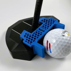 May include: A golf putting alignment tool with a black base and a blue alignment guide. A white golf ball with red and blue stripes is positioned next to the guide. The golf club is inserted into the base.