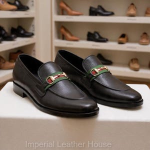 May include: A pair of black leather loafers with a gold and green accent strap. The shoes are displayed on a white surface, with other shoes visible on shelves in the background. The text "Imperial Leather House" is at the bottom.