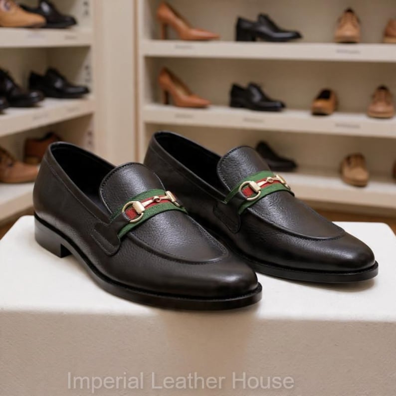 May include: A pair of black leather loafers with a gold and green accent strap. The shoes are displayed on a white surface, with other shoes visible on shelves in the background. The text "Imperial Leather House" is at the bottom.