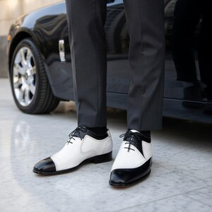 May include: Two-tone black and white leather dress shoes with black laces. The shoes have a classic design with a black toe cap and heel, and a white upper. The shoes are worn with dark gray trousers.