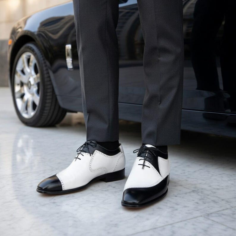 May include: Two-tone black and white leather dress shoes with black laces. The shoes have a classic design with a black toe cap and heel, and a white upper. The shoes are worn with dark gray trousers.