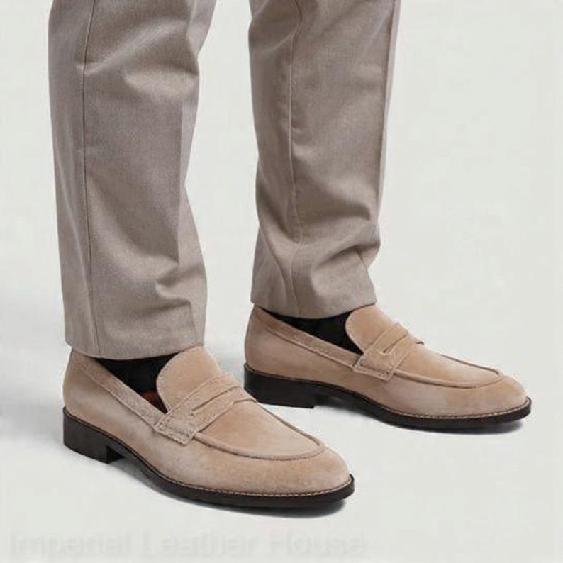 May include: Tan suede penny loafers with a dark brown sole. The shoes are worn with light brown trousers and black socks. The loafers have a classic design with a strap across the top.