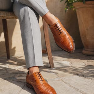 Puede incluir: Un par de zapatos oxford de cuero color canela con textura trenzada en la puntera y los paneles laterales, detalles de cuero liso en el cierre de cordones, cordones marr&oacute;n oscuro y tac&oacute;n bajo, usados con pantalones gris claro.