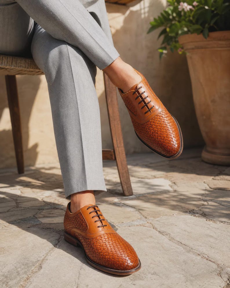 Puede incluir: Un par de zapatos oxford de cuero color canela con textura trenzada en la puntera y los paneles laterales, detalles de cuero liso en el cierre de cordones, cordones marr&oacute;n oscuro y tac&oacute;n bajo, usados con pantalones gris claro.