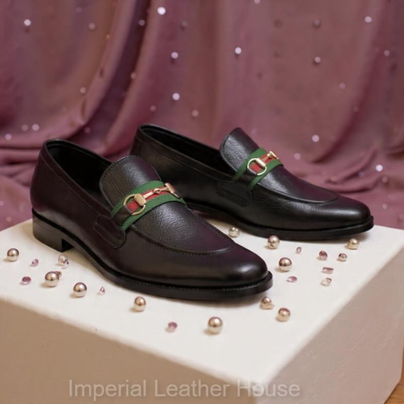 May include: A pair of black leather loafers with a decorative green and red strap. The shoes are displayed on a white surface with scattered metallic beads. The text "Imperial Leather House" is at the bottom.
