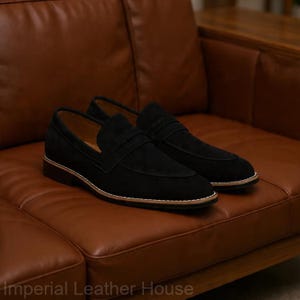 May include: A pair of black suede loafers with a penny strap detail, set on a brown leather couch. The shoes have a light-colored sole and a dark brown heel.