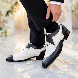 May include: Two-tone black and white dress shoes with a classic design. The shoes feature a black toe cap, heel, and trim, with white panels and decorative perforations. The shoes are laced up and appear to be made of leather. The image shows the shoes from a low angle.