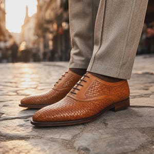 Puede incluir: Un par de zapatos oxford de color tostado con textura de cuero trenzado, cordones marr&oacute;n oscuro y tac&oacute;n bajo. Los zapatos se llevan con pantalones de color marr&oacute;n claro con pliegue, mostrando un dise&ntilde;o cl&aacute;sico de cordones con puntera lisa.