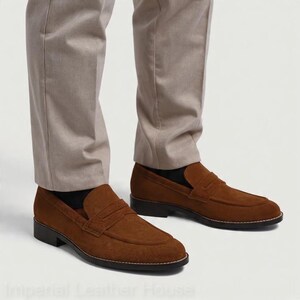 May include: Brown suede penny loafers with a classic design. The shoes have a rounded toe and a low heel. The loafers are paired with light tan trousers. The shoes are a stylish choice for casual or semi-formal wear.