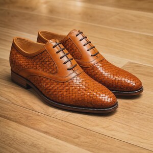 Puede incluir: Un par de zapatos oxford de cuero color canela con una textura tejida en la parte superior, paneles de cuero liso en el cierre de cordones, cordones marr&oacute;n oscuro y un tac&oacute;n bajo. Los zapatos tienen una punta redondeada y una silueta estructurada.