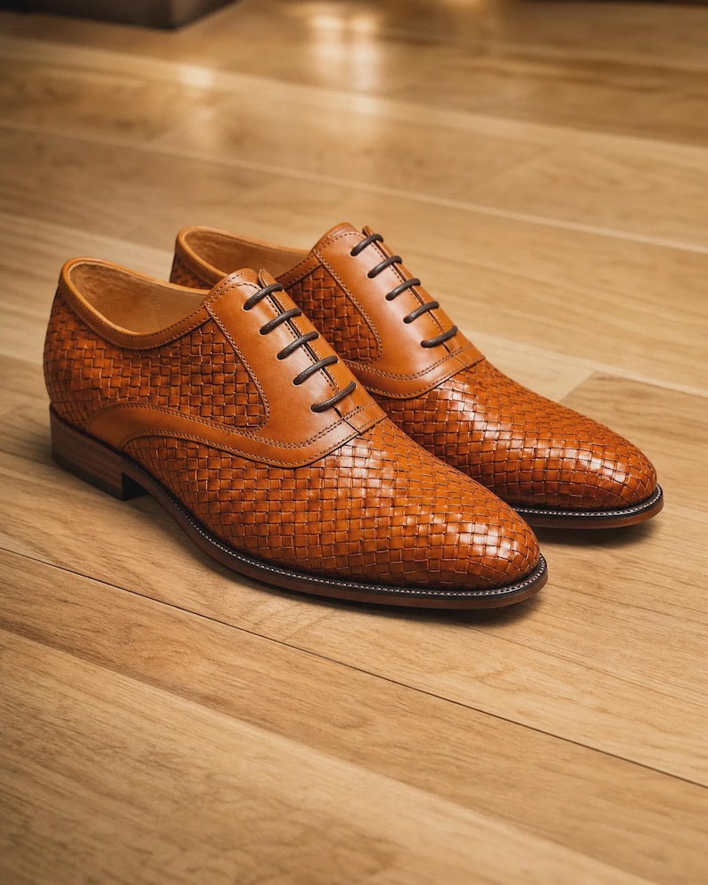 Puede incluir: Un par de zapatos oxford de cuero color canela con una textura tejida en la parte superior, paneles de cuero liso en el cierre de cordones, cordones marr&oacute;n oscuro y un tac&oacute;n bajo. Los zapatos tienen una punta redondeada y una silueta estructurada.