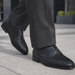 Puede incluir: Un par de zapatos oxford negros con una textura de cuero trenzado en la punta y los paneles laterales, complementados con detalles de cuero liso en el cierre de cordones y el tal&oacute;n. Los zapatos tienen un tac&oacute;n bajo y pantalones gris oscuro.