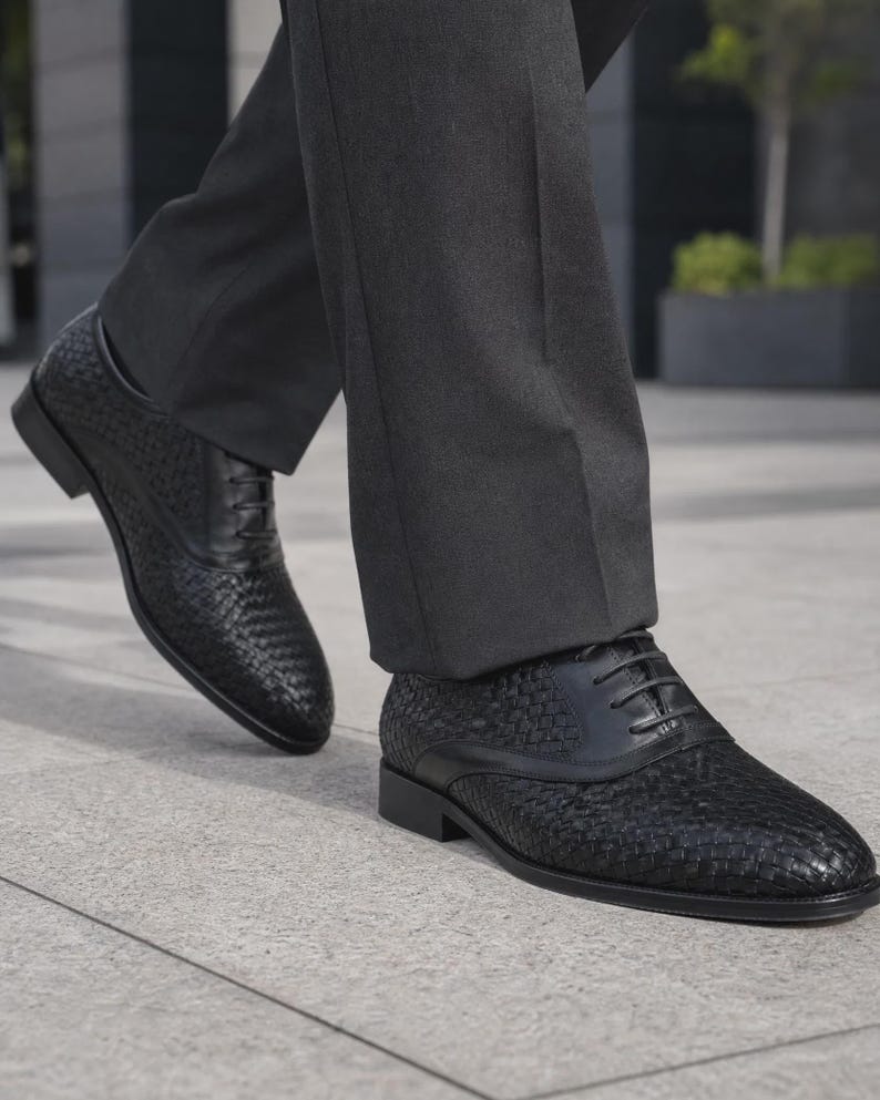 Puede incluir: Un par de zapatos oxford negros con una textura de cuero trenzado en la punta y los paneles laterales, complementados con detalles de cuero liso en el cierre de cordones y el tal&oacute;n. Los zapatos tienen un tac&oacute;n bajo y pantalones gris oscuro.