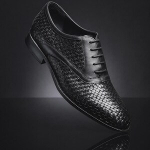 Puede incluir: Un zapato oxford de cuero negro con una textura tejida en la parte superior, un panel de cordones de cuero liso con cordones negros a juego, una punta redondeada y un tac&oacute;n bajo apilado. El zapato se presenta en &aacute;ngulo.