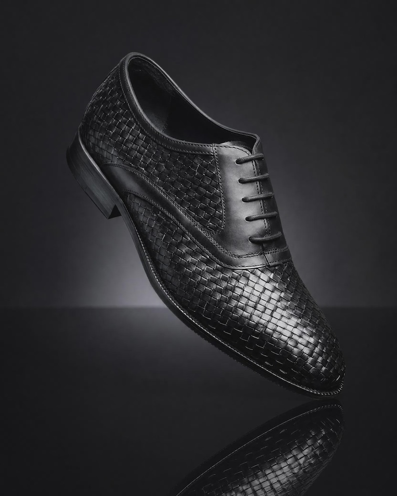 Puede incluir: Un zapato oxford de cuero negro con una textura tejida en la parte superior, un panel de cordones de cuero liso con cordones negros a juego, una punta redondeada y un tac&oacute;n bajo apilado. El zapato se presenta en &aacute;ngulo.