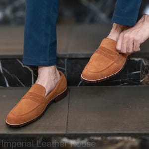 May include: A pair of brown suede loafers with a penny strap detail. The shoes have a dark brown sole and a white stitched edge. The person is wearing blue pants. The text "Imperial Leather House" is at the bottom of the image.