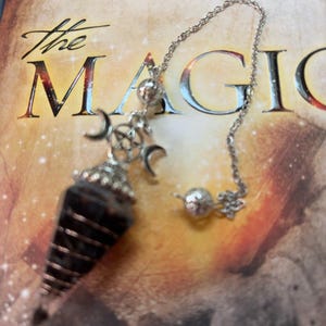 May include: A dark, faceted, cone-shaped crystal pendulum wrapped in silver-toned wire. It features a silver-toned metal cap with a triple moon and pentagram symbol, attached to a silver-toned chain with two small, textured, spherical metal beads.
