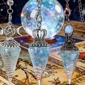 May include: Three crystal pendulums with silver and copper accents. Each pendulum features a blue-toned crystal encased in a decorative metal setting. One has a triple moon design, another a crown, and the third a small crystal sphere.