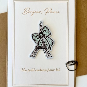 May include: Greeting card with a brown border and the text "Bonjour, Paris" in cursive. An embroidered Eiffel Tower with a bow is attached. The card also has the text "Un petit cadeau pour toi." A small, black, wire-shaped design is on the right.
