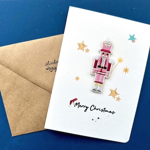 Peut inclure: Une carte de Noël blanche avec un motif de casse-noisette rose et l'inscription "Merry Christmas". La carte est décorée d'étoiles dorées et d'un petit bonnet de Père Noël rouge. Une enveloppe marron avec le mot "thinking" est à gauche.