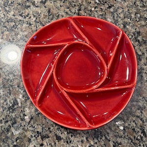 May include: A circular red ceramic serving dish featuring a glossy finish and a segmented design. The plate includes a central triangular compartment surrounded by five distinct, radiating wedge-shaped sections separated by raised dividers.