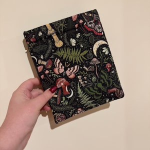 May include: A black pouch with a whimsical botanical print featuring mushrooms, moths, ferns, and crescent moons. The design includes shades of red, pink, green, and cream. The pouch is held in a hand.