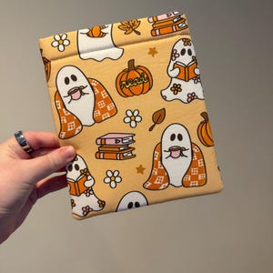 May include: A rectangular fabric sleeve with a light orange background featuring a repeating pattern of white ghosts, orange pumpkins with the word read, stacks of books, and white flowers. The ghosts are depicted drinking from mugs.