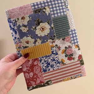 May include: A patchwork fabric book cover featuring a variety of floral and patterned fabrics in shades of blue, pink, yellow, and red. The cover is rectangular and appears to be handmade, with a mix of floral and gingham patterns.