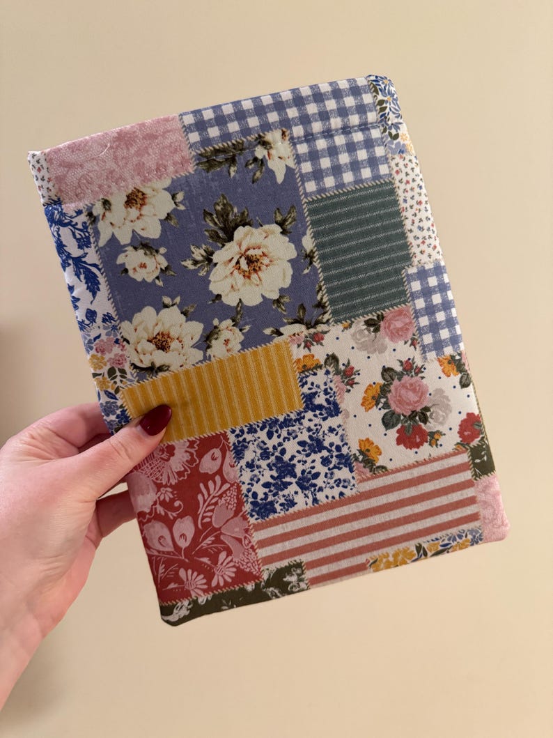 K&ouml;nnte beinhalten: Ein Patchwork-Stoff-Buchumschlag mit einer Vielzahl von Stoffen mit Blumen- und Mustermotiven in Blau-, Rosa-, Gelb- und Rott&ouml;nen. Der Umschlag ist rechteckig und scheint handgefertigt zu sein, mit Blumen- und Vichy-Mustern.
