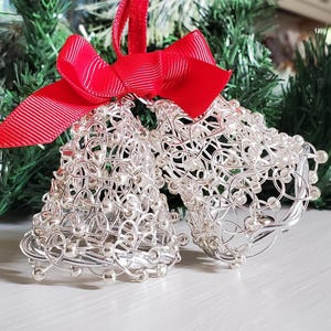 May include: Two silver wire bell ornaments with small silver beads, connected by a red ribbon bow. The bells are in front of a green Christmas tree. A festive holiday decoration.