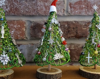 May include: Three handmade Christmas trees crafted from green wire, adorned with beads and charms. Each tree sits on a wooden base. One tree has a Santa hat, and another has a "Merry Christmas" sign.