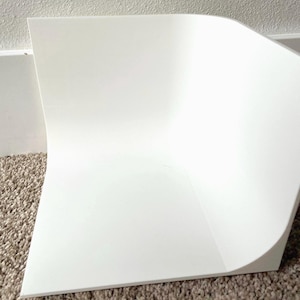 May include: A white, curved, rectangular backdrop for product photography. The backdrop is made of a thick, opaque material and is positioned on a neutral-colored carpet. The curved design creates a seamless background for showcasing items.