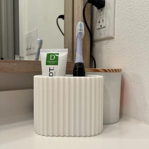 May include: A white, ribbed toothbrush holder with a toothbrush and toothpaste tube. The toothpaste tube has green and white text. A white ceramic container with a wooden lid is to the right. The items are on a white countertop.