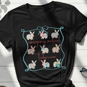 May include: Black t-shirt featuring a design with nine watercolor bunnies inside a teal frame. The text "emergency medical technician" is written in cursive. A coffee cup and glasses are also visible.