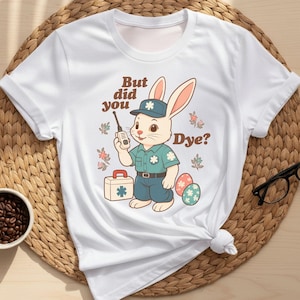 May include: White t-shirt featuring a cartoon bunny in a uniform holding a walkie-talkie, with the text "But did you Dye?" Easter-themed design includes eggs and a first aid kit. The shirt is styled on a woven mat.