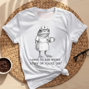 Op de afbeelding: Wit t-shirt met een gedetailleerde illustratie van een kikker in een verpleegstersuniform, met een mok. De tekst "I HAVE TO ASK WEIRD STUFF OR YOU'LL DIE" staat eronder. Het shirt is onderaan geknoopt.