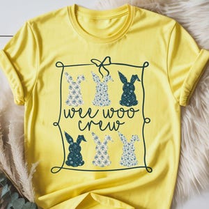 May include: A yellow t-shirt with a crew neck and rolled-up sleeves. The shirt features a graphic of six bunny silhouettes with floral patterns and the text "wee woo crew" in a black frame. The word "cornsilk" is at the bottom.