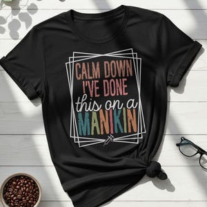May include: Black t-shirt with the text "CALM DOWN I'VE DONE this on a MANIKIN" in a colorful, retro design. The shirt features a syringe graphic and is knotted at the waist. A pair of glasses and a bowl of coffee beans are also visible.