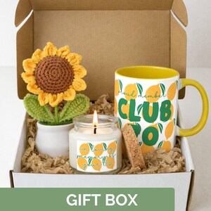 Op de afbeelding: Een geschenkdoos met een gehaakte zonnebloem, een kaars met een citroenontwerp, een mok met een citroenontwerp en de tekst "Club Yo", en een kurk. De doos heeft de tekst "Gift Box" aan de onderkant.