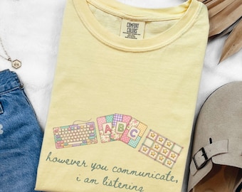 Puede incluir: Camiseta amarilla clara con un diseño de teclado, tarjetas ABC y una cuadrícula de estrellas. El texto "however you communicate, i am listening" está impreso debajo. La camiseta es de material suave.