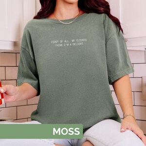 Puede incluir: Camiseta verde musgo con el texto blanco "FIRST OF ALL, MY CLIENTS THINK I'M A DELIGHT". La camiseta tiene un corte holgado y mangas cortas. La palabra "MOSS" se muestra en un rect&aacute;ngulo verde en la parte inferior.