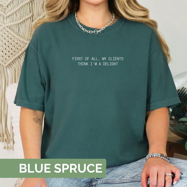 Puede incluir: Camiseta azul abeto con el texto blanco "FIRST OF ALL, MY CLIENTS THINK I'M A DELIGHT". La camiseta tiene cuello redondo y mangas cortas. Las palabras "BLUE SPRUCE" est&aacute;n en la parte inferior de la imagen.